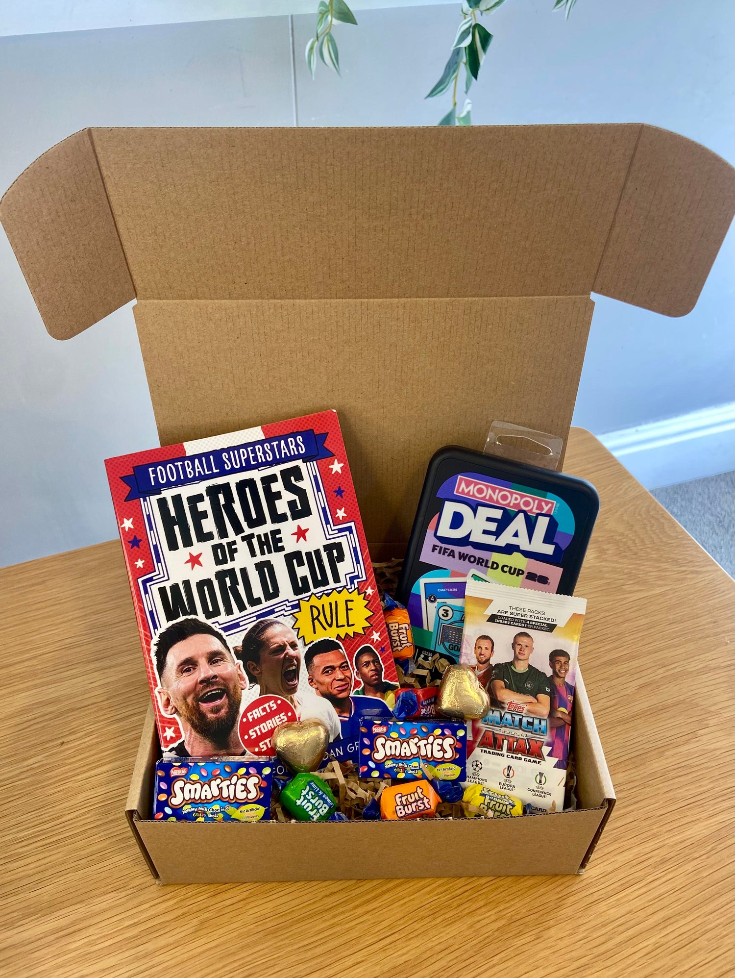 A cardboard gift box open on a wooden table, filled with a red 'Heroes of the World Cup' football magazine, a black 'Monopoly Deal FIFA World Cup' tin, and assorted small snacks and chocolates.