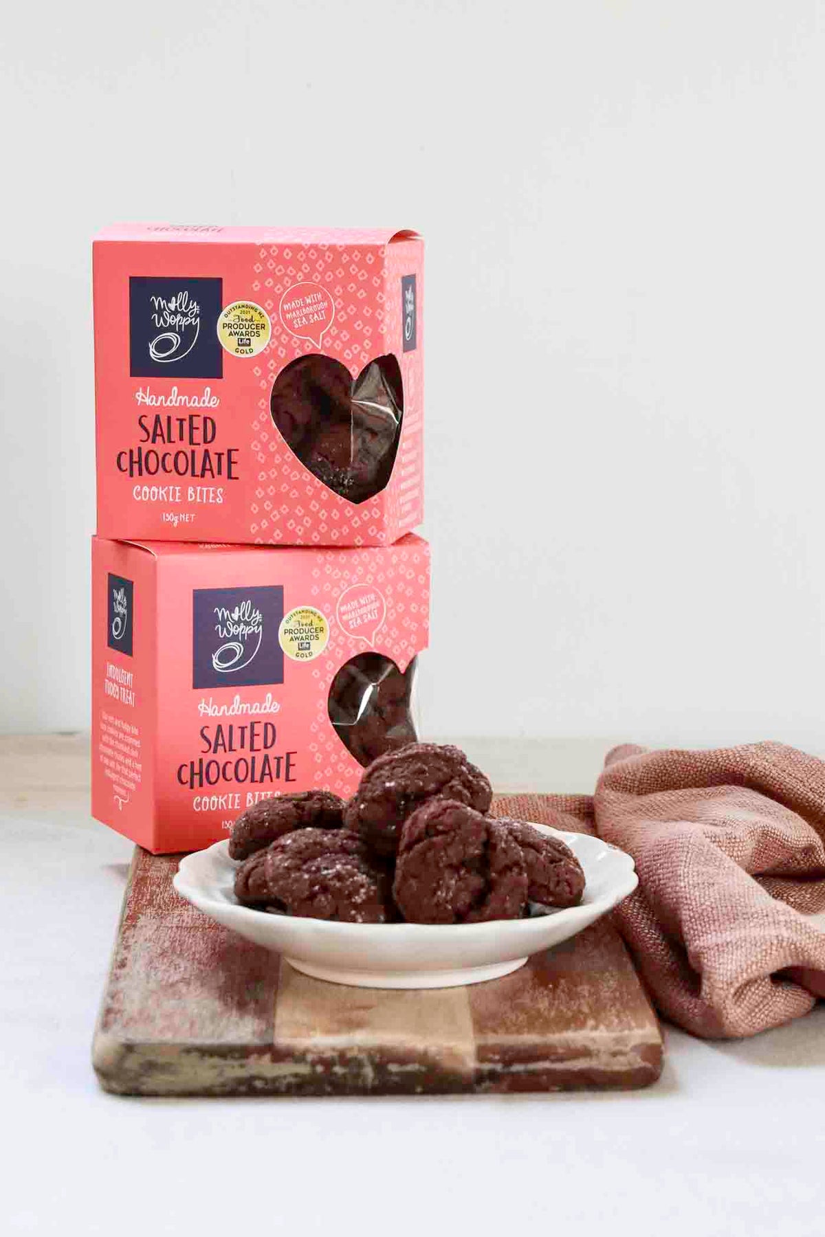 Two pink boxes of Truffa Lifestyle Handmade Salted Chocolate Cookie Bites stacked on a wooden cutting board, with a white plate of chocolate cookie bites and a dusty pink cloth on a white background.