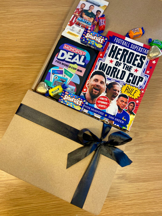 A brown kraft gift box tied with a dark ribbon, opened to reveal a curated football-themed gift set including a Monopoly Deal card game, a Heroes of the World Cup magazine, and Snickersa's chocolate bars, on a wooden tabletop.