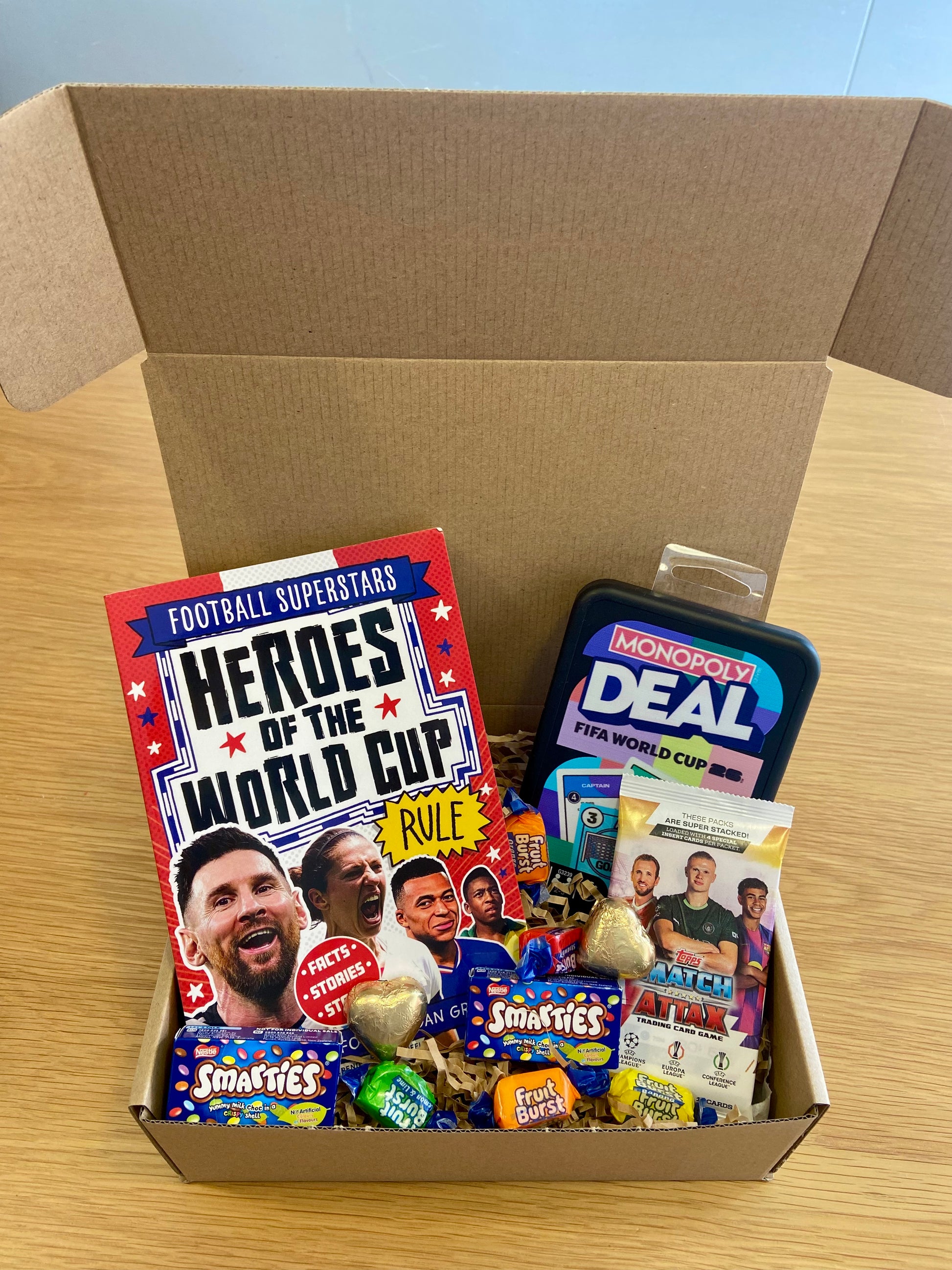 A cardboard gift box filled with football-themed items, including a book titled 'Heroes of the World Cup', a Monopoly Deal tin, and assorted branded candy and snack packs, set on a light wood tabletop.