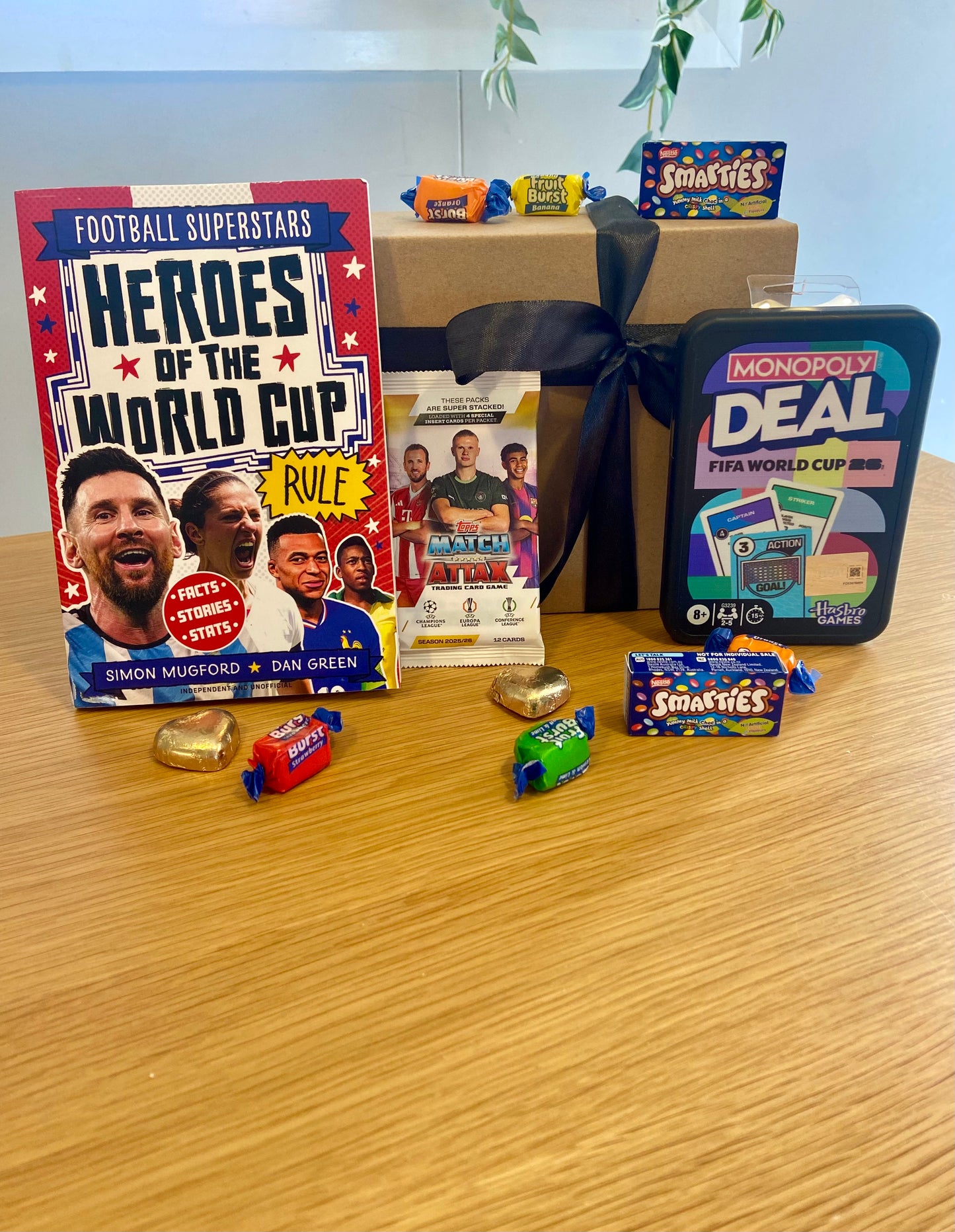 A curated gift set arranged on a light wood tabletop, featuring a 'Heroes of the World Cup' football book, a Monopoly Deal tin, a small SMRTS card pack, several wrapped chocolates, and a brown gift box tied with a black ribbon.
