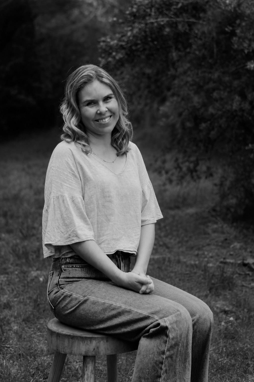 A woman sitting on a wooden stool outdoors, wearing a light-colored short-sleeve top and jeans, captured in a black and white photograph.