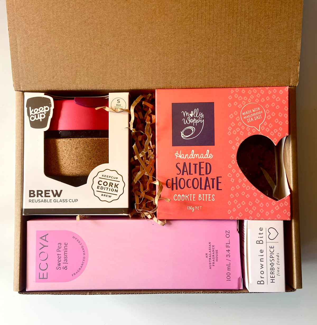 An open cardboard gift box containing multiple packaged items: a reusable glass cup with a pink lid and cork insert from Keep Cup, a pink box of Ecolya Sweet Tea in Syrup, a pink box of Brownie Bite Herbspice, and a red box of Molly's Waynes Handmade Salted Chocolate Cookie Bites.