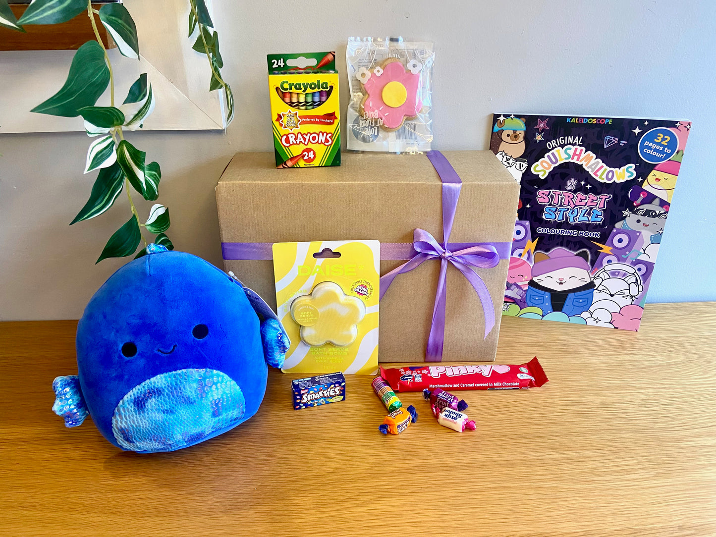 A Kraft gift box with a purple with a purple ribbon for kids birthday and gifts. Inside are assorted treats and toys including a Crayola crayons, Daise Bath bomb, Squishmallow toy and colouring book, pinky bar and more.