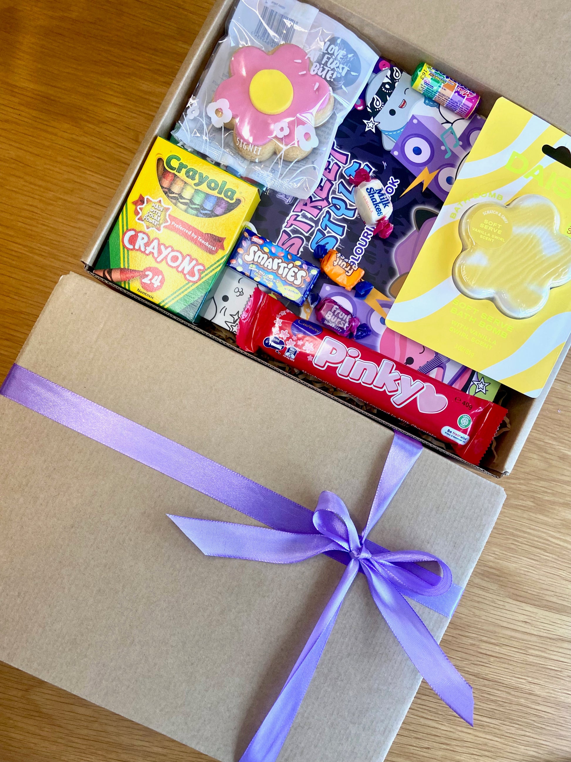 A Kraft gift box with a purple with a purple ribbon. Inside are assorted treats and toys including a Crayola crayons, Daise Bath bomb, Squishmallow toy and colouring book, pinky bar and more.
