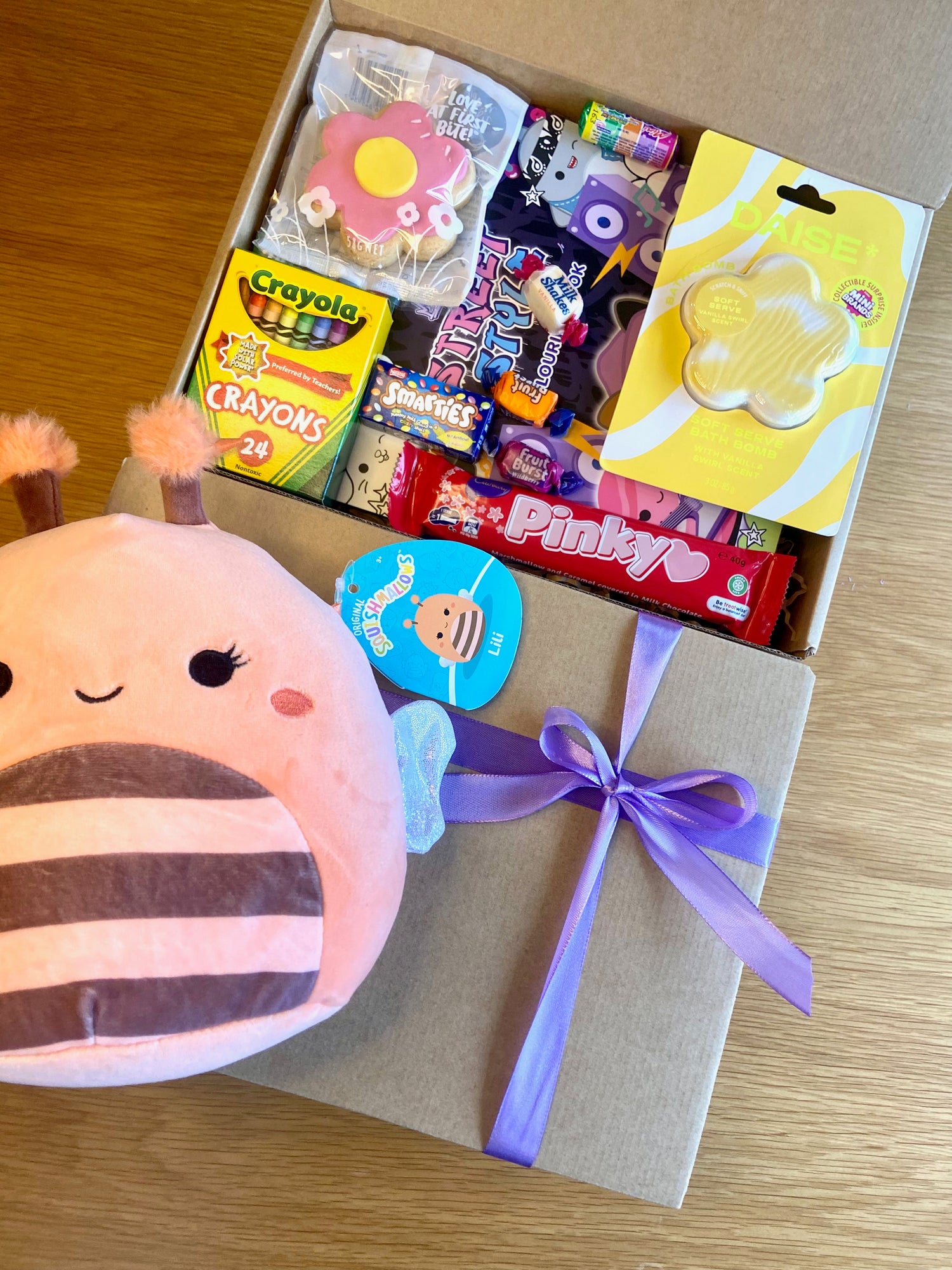 A Kraft gift box with a purple with a purple ribbon for kids birthday and gifts. Inside are assorted treats and toys including a Crayola crayons, Daise Bath bomb, Squishmallow toy and colouring book, pinky bar and more.