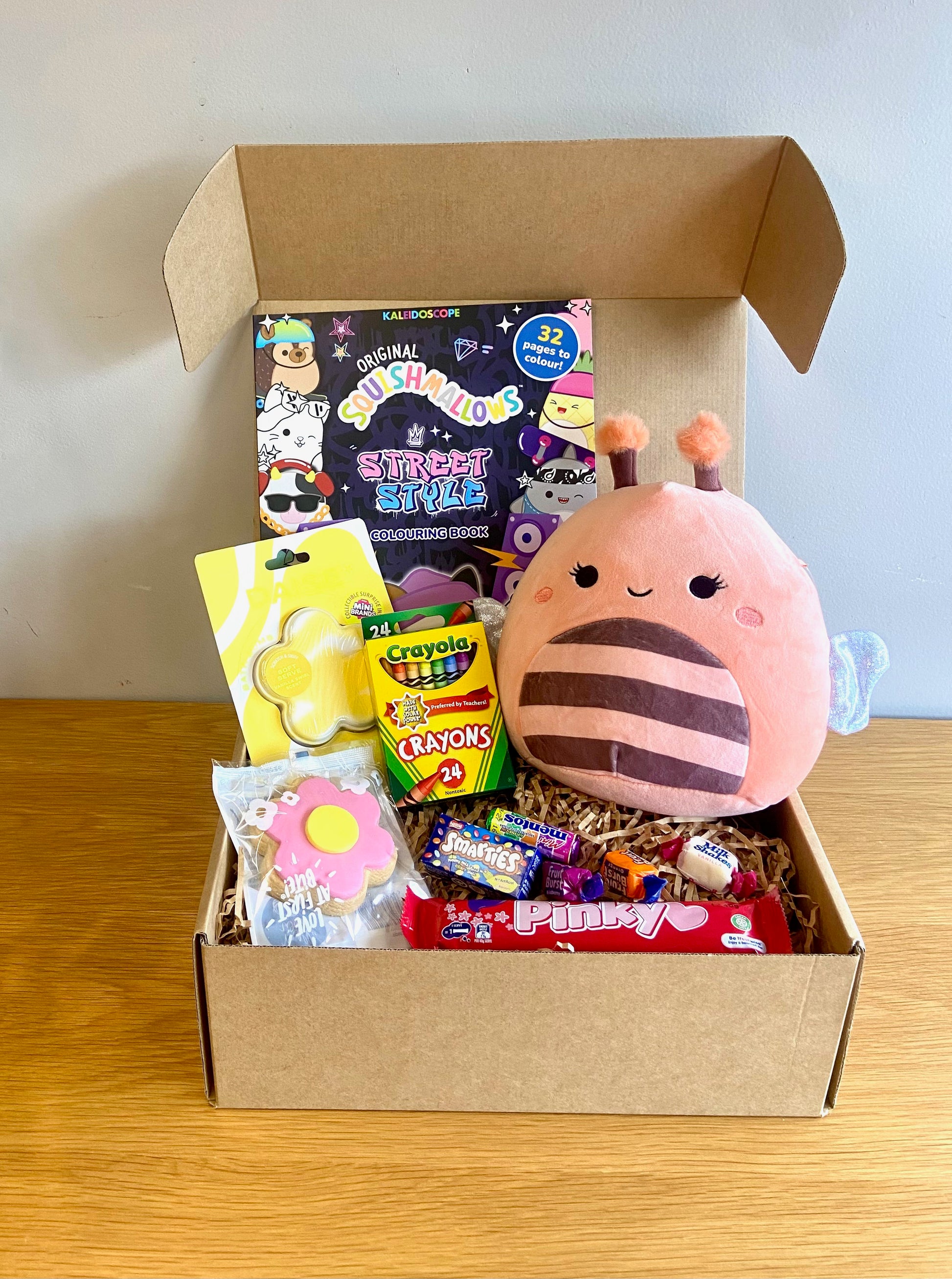 A Kraft gift box with a purple with a purple ribbon for kids birthday and gifts. Inside are assorted treats and toys including a Crayola crayons, Daise Bath bomb, Squishmallow toy and colouring book, pinky bar and more.