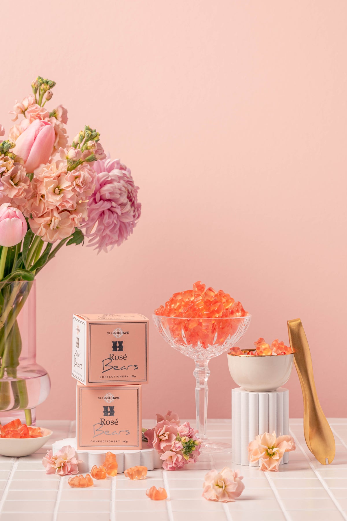 Pink rose-bean gel products displayed with pink packaging boxes, a glass bowl of orange-pink gel, flowers, and a gold spoon on a pink background.
